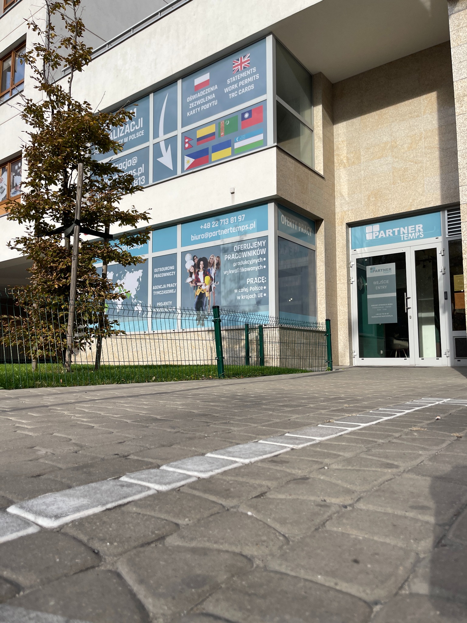 Budynek firmy Partner Temps z widocznymi oknami z reklamami usług, flagami różnych krajów, oraz informacjami kontaktowymi. Przed budynkiem chodnik z kostki brukowej z namalowaną białą linią.
