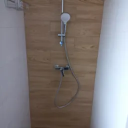 Nowoczesny prysznic z panelami imitującymi drewno i chromowaną armaturą. Widoczna kratka odpływowa w podłodze. Minimalistyczny design i jasne kolory.