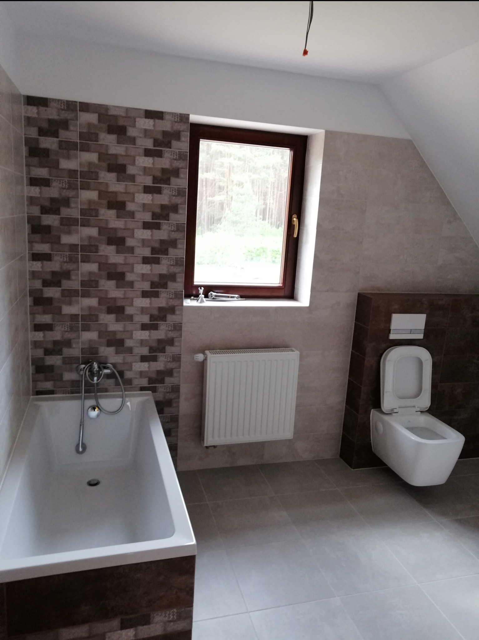 Nowoczesna łazienka na poddaszu z brązowymi akcentami: mozaika, zabudowa toalety, rama okna. Biała wanna, grzejnik i toaleta pod oknem. Szare płytki podłogowe.
