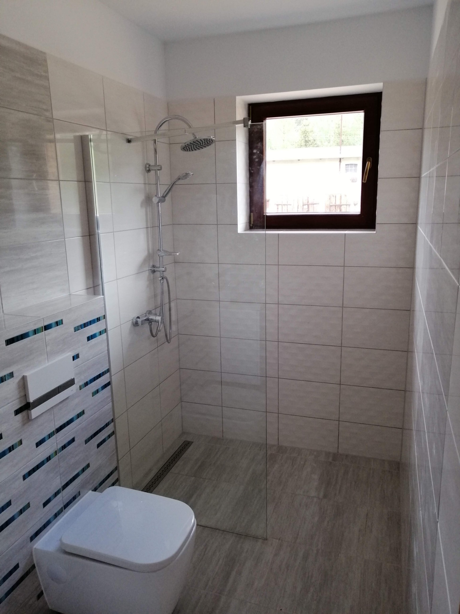 Nowoczesna łazienka z kabiną typu walk-in, wykończona płytkami w odcieniach szarości i bieli. Uwagę zwracają dekoracyjne, niebieskie akcenty na ścianie z toaletą.