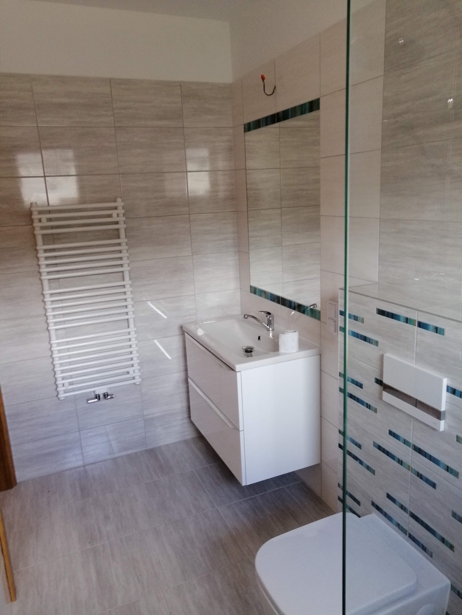 Jasna łazienka po remoncie: płytki w odcieniach szarości, biała szafka z umywalką, grzejnik drabinkowy i lustro z dekoracyjnym paskiem. Widoczna kabina prysznicowa.