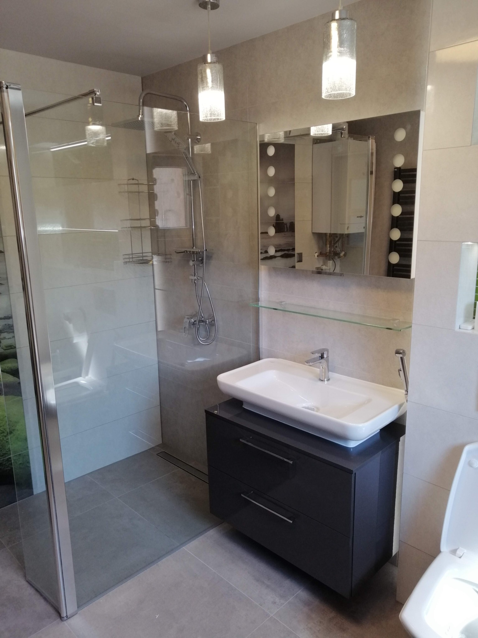 Nowoczesna łazienka w szarościach z kabiną prysznicową typu walk-in, szafką pod umywalką i lustrem z oświetleniem. Minimalistyczny design z wiszącymi lampami.