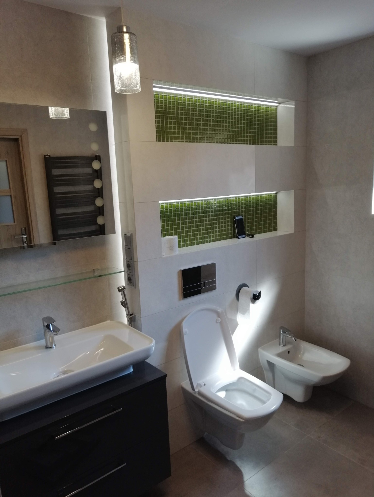 Nowoczesna łazienka z białą ceramiką, podświetlanymi niszami z zieloną mozaiką i wiszącą lampą. Umywalka nablatowa na ciemnej szafce.