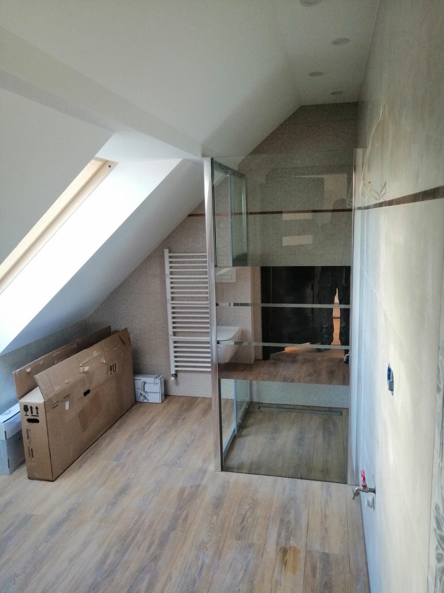 Wykończona łazienka na poddaszu z kabiną prysznicową, grzejnikiem drabinkowym i drewnopodobną podłogą. Widoczne kartony po sprzęcie.