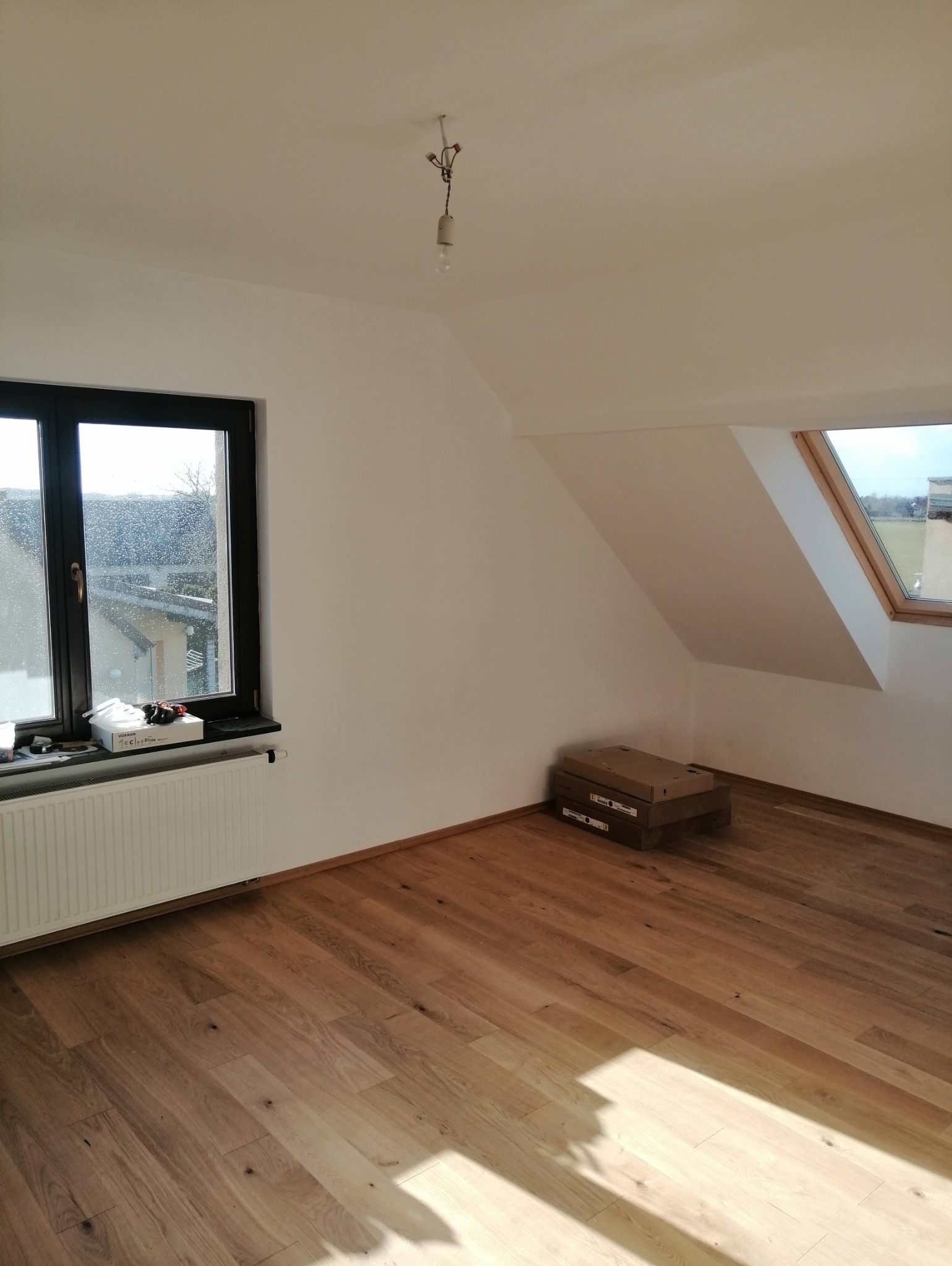 Jasne, nowo wykończone poddasze z drewnianą podłogą, oknem dachowym i grzejnikiem pod oknem. Na podłodze leżą kartony.