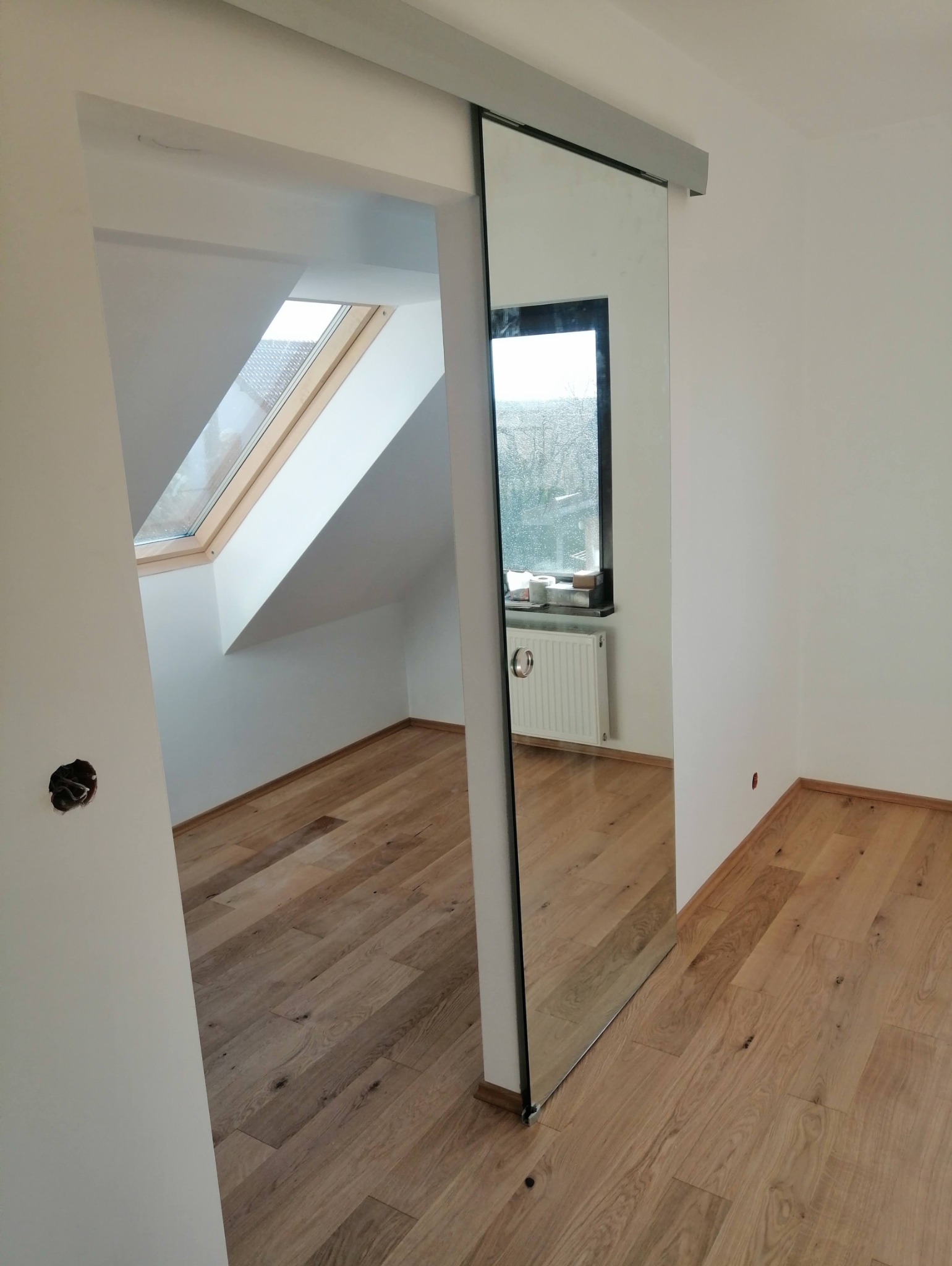 Montaż drzwi przesuwnych z lustrem, widok na poddasze z oknem dachowym i drewnianą podłogą. Nowoczesne wykończenie wnętrza, białe ściany.