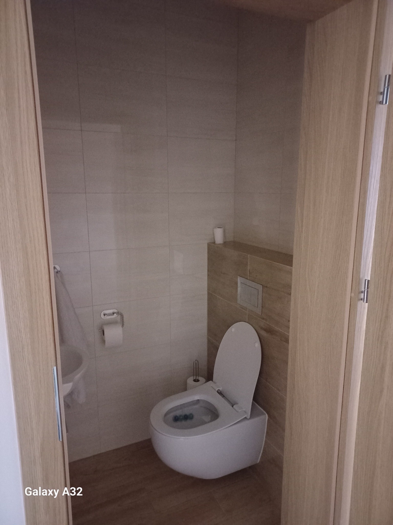 Nowoczesna łazienka z podwieszaną toaletą i beżowymi płytkami. Widoczne drewniane drzwi i minimalistyczne akcesoria. Stonowana kolorystyka i proste linie.