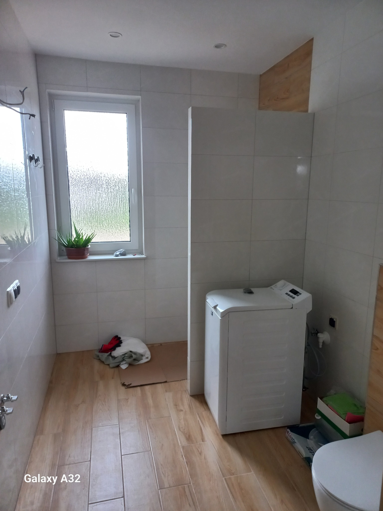 Jasna łazienka z drewnopodobną podłogą i białymi płytkami. Widoczna pralka, okno z rośliną i fragment toalety. Ułożona podłoga i wykończone ściany.