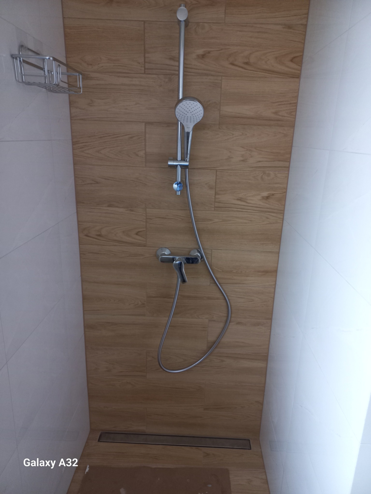 Nowoczesny prysznic z panelami imitującymi drewno i chromowaną armaturą. Widoczna kratka odpływowa w podłodze. Minimalistyczny design i jasne kolory.