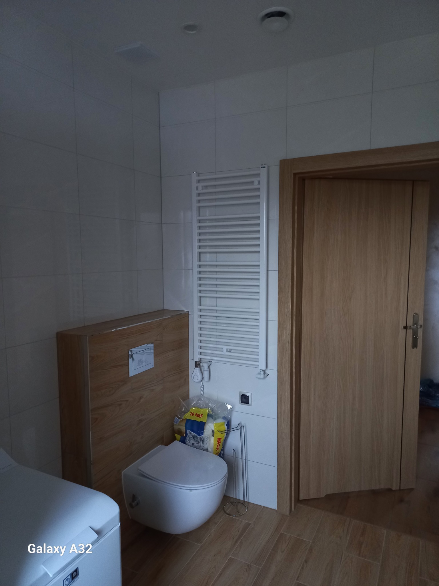 Wykończona łazienka w jasnych płytkach z drewnopodobną podłogą. Widoczny podwieszany sedes, grzejnik drabinkowy i dębowe drzwi. Ujęcie z niskiego kąta.