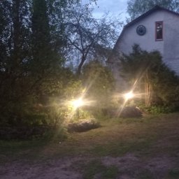 DZMITRY KUPREYEU - Oświetlenie zewnętrzne posesji z dwoma niskimi lampami ogrodowymi o ciepłej barwie, podświetlającymi roślinność przed domem z dekoracyjnym kołem na szczycie