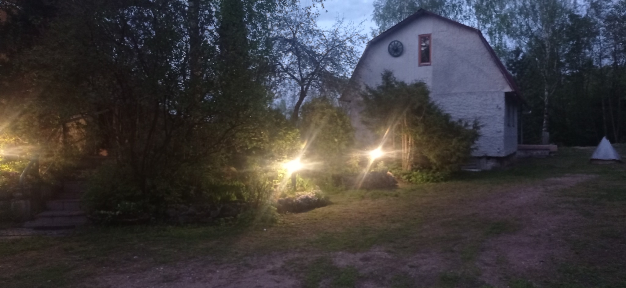 Oświetlenie zewnętrzne posesji z dwoma niskimi lampami ogrodowymi o ciepłej barwie, podświetlającymi roślinność przed domem z dekoracyjnym kołem na szczycie