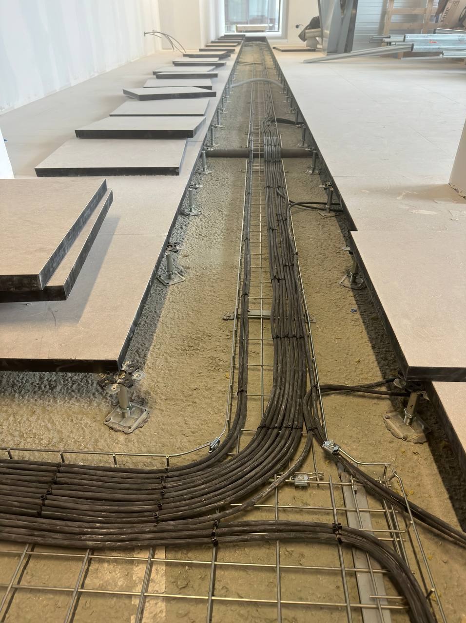 Montaż podłogi podniesionej z widocznymi trasami kablowymi w korycie pod podłogą. Kable ułożone równolegle w metalowej siatce. Panele podłogowe częściowo ułożone na regulowanych wspornikach.