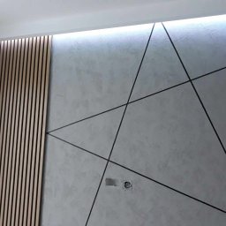 Jarosław Siemiński - Nowoczesna ściana z dekoracyjną strukturą, czarnymi liniami tworzącymi geometryczny wzór i drewnianymi panelami lamelowymi po lewej stronie, oświetlenie LED wzdłuż sufitu, widoczne gniazdko...