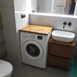 Nowoczesna łazienka z pralką zabudowaną pod drewnianym blatem, umywalką nablatową i podświetlanym lustrem.