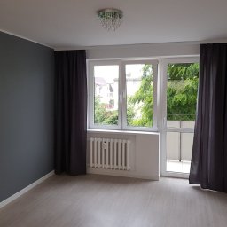 Jarosław Siemiński - Pusty pokój po remoncie z szarą ścianą, jasnymi panelami podłogowymi, białym grzejnikiem pod oknem balkonowym i ciemnymi zasłonami.