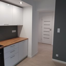 Jarosław Siemiński - Nowoczesna kuchnia z białymi szafkami, drewnianym blatem i ciemnoszarą ścianą, widok na białe drzwi z poziomymi czarnymi paskami.