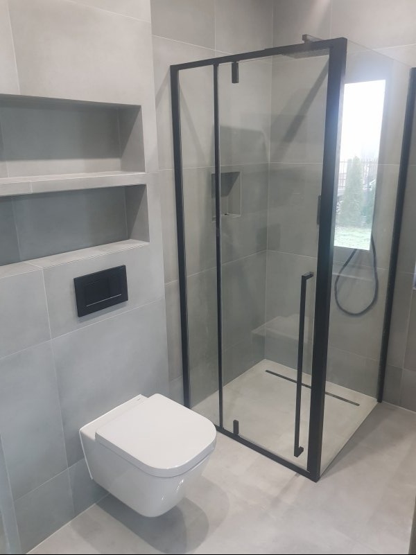 Nowoczesna łazienka w odcieniach szarości z kabiną prysznicową z czarnymi profilami, podwieszaną toaletą i wnękami w ścianie wyłożonymi płytkami.
