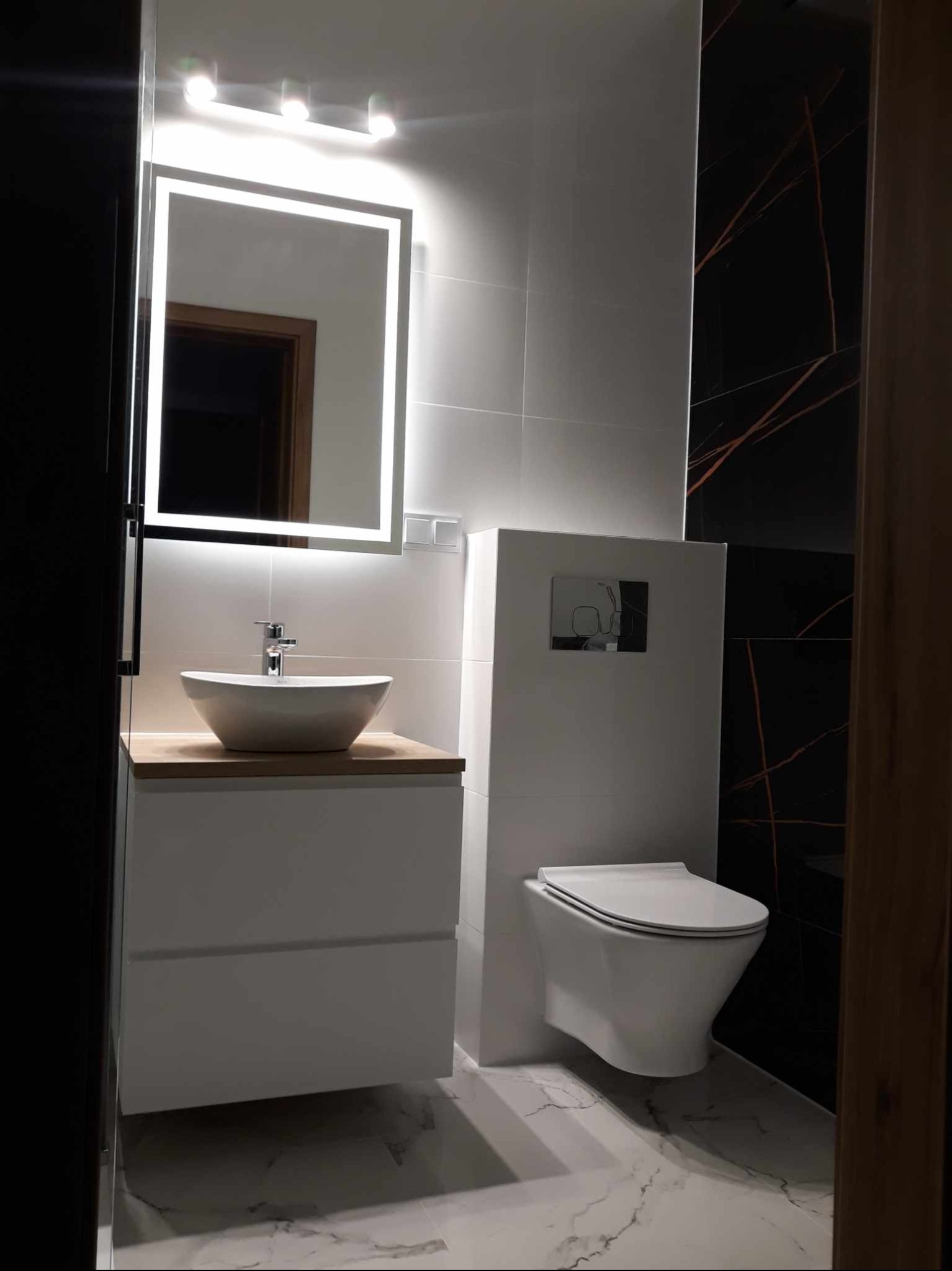 Nowoczesna łazienka z umywalką nablatową na drewnianym blacie, podświetlanym lustrem, podwieszaną toaletą i czarno-białymi płytkami imitującymi marmur.