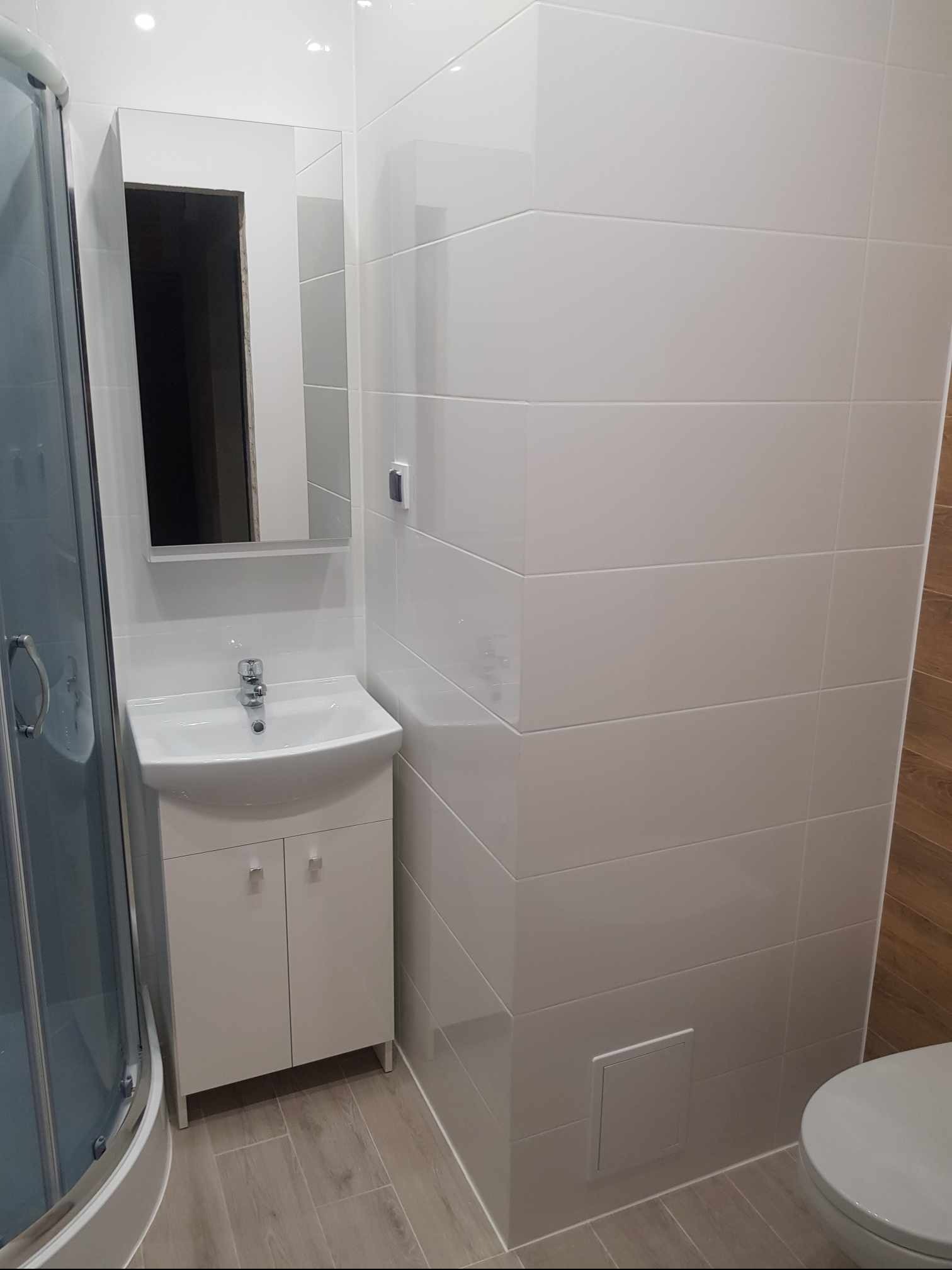 Jasna łazienka po remoncie: białe, poziome płytki ścienne, umywalka z szafką, lustro z oświetleniem, kabina prysznicowa z przezroczystymi drzwiami, drewnopodobne płytki podłogowe.