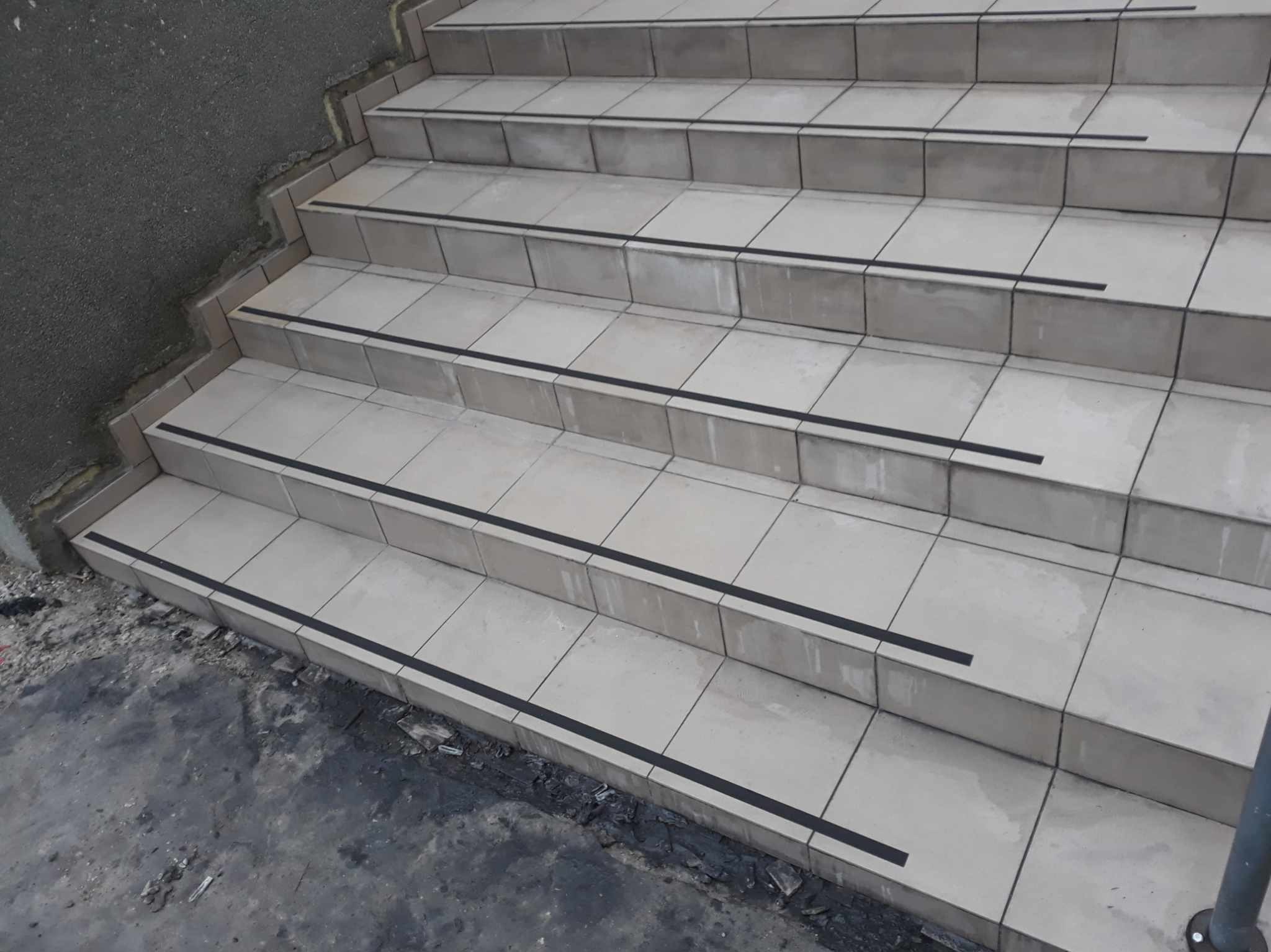 Wyłożone jasnymi płytkami schody z czarnymi antypoślizgowymi paskami, widoczne ślady po remoncie na podłodze.