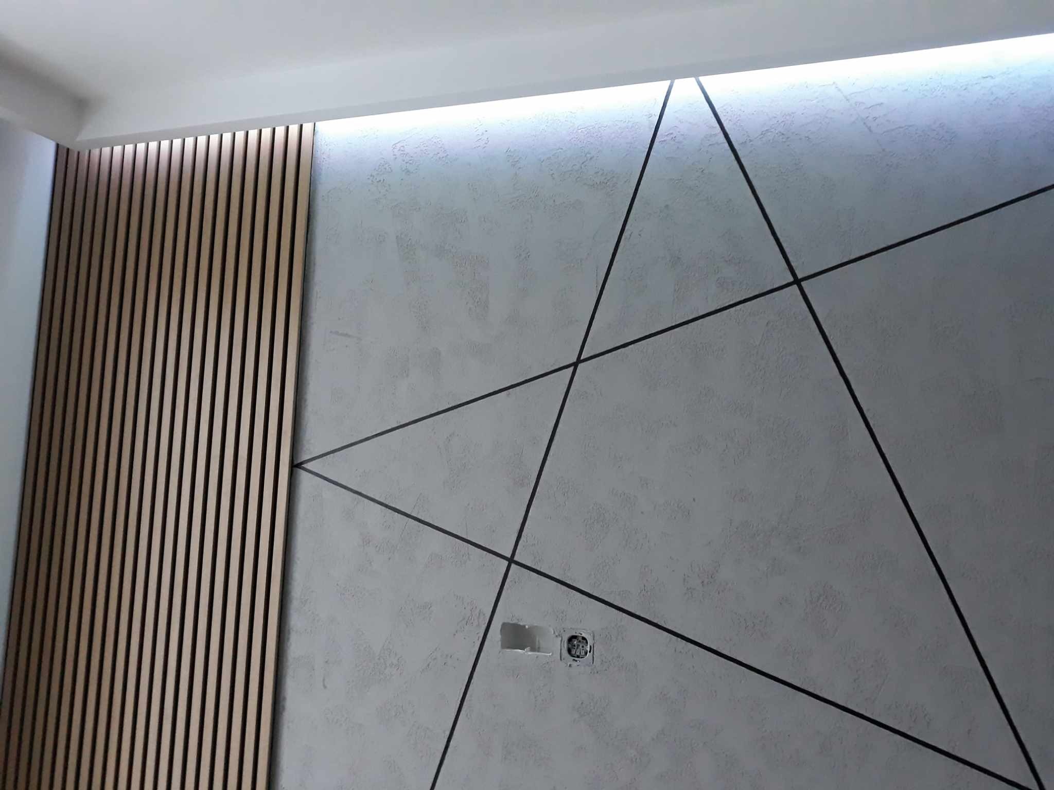 Nowoczesna ściana z dekoracyjną strukturą, czarnymi liniami tworzącymi geometryczny wzór i drewnianymi lamelami po lewej stronie, oświetlona od góry, z widocznym gniazdkiem elektrycznym.