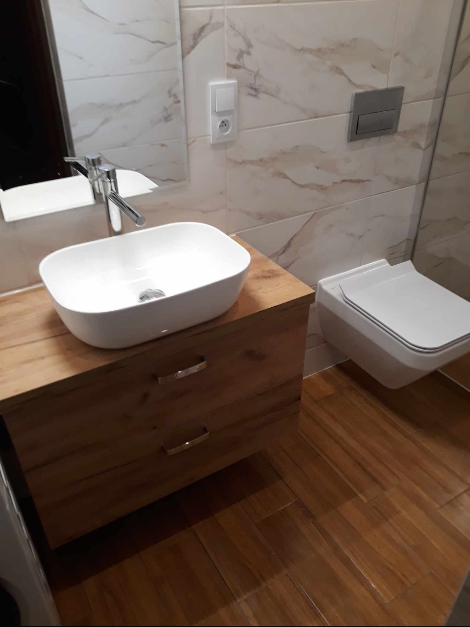 Nowoczesna łazienka z podwieszaną toaletą, umywalką nablatową na szafce z drewnopodobnym frontem i marmurkowymi płytkami ściennymi.