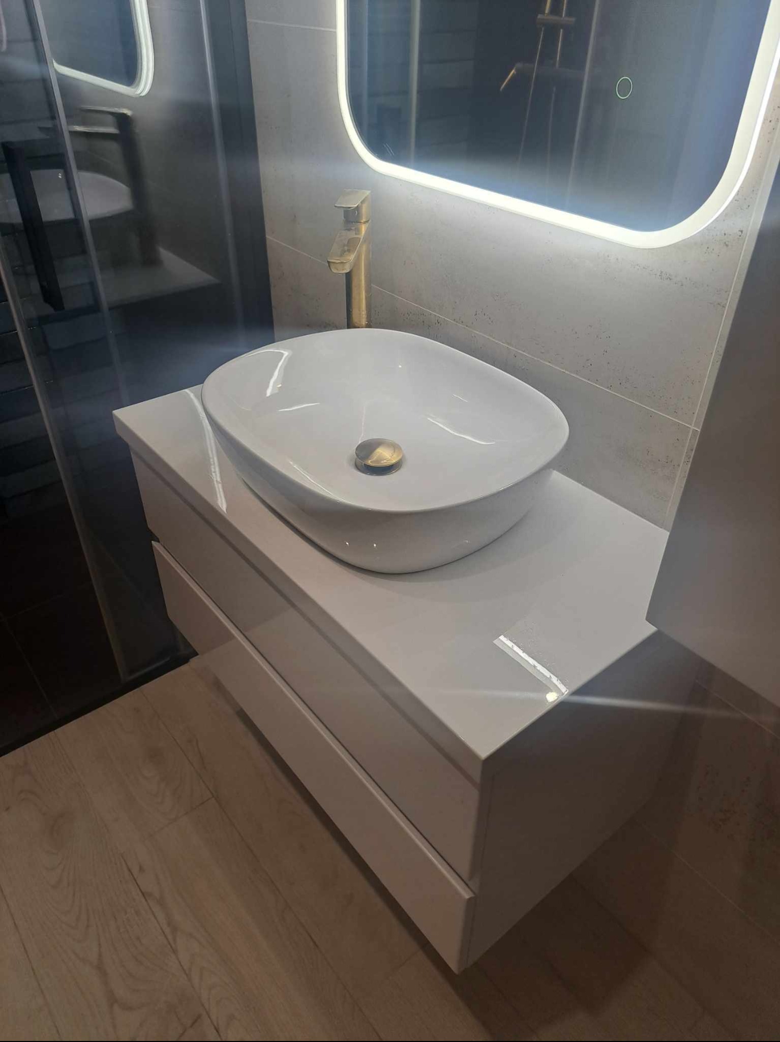 Nowoczesna łazienka z białą szafką pod umywalkę, owalną umywalką nablatową i złotą armaturą, oświetlone lustro LED, szare płytki na ścianie, drewniana podłoga.