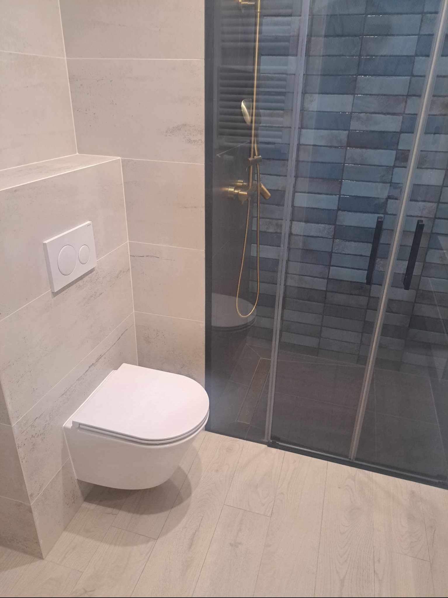 Nowoczesna łazienka z podwieszaną toaletą, jasnymi płytkami imitującymi kamień i kabiną prysznicową z niebiesko-szarymi płytkami w cegiełkę oraz złotą armaturą.