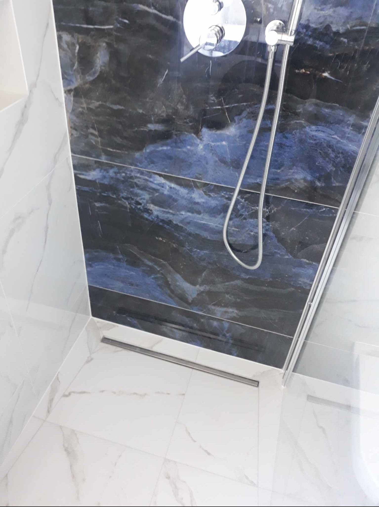 Kabina prysznicowa z odpływem liniowym, wykończona białymi płytkami imitującymi marmur na podłodze i ścianach, oraz płytkami w odcieniach granatu i szarości z efektem marmuru na ścianie prysznica.
