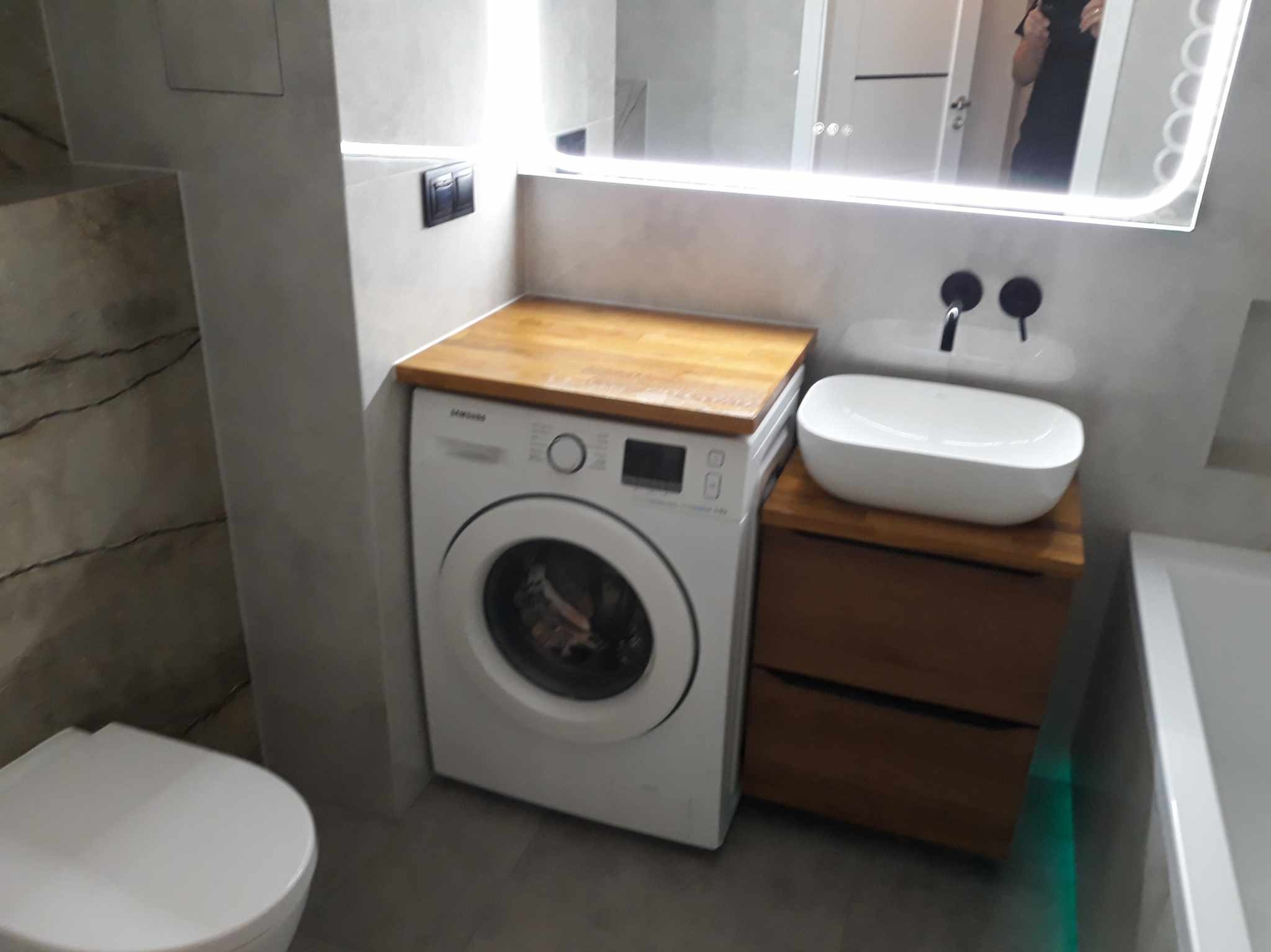 Nowoczesna łazienka z pralką zabudowaną pod drewnianym blatem, umywalką nablatową i podświetlanym lustrem.