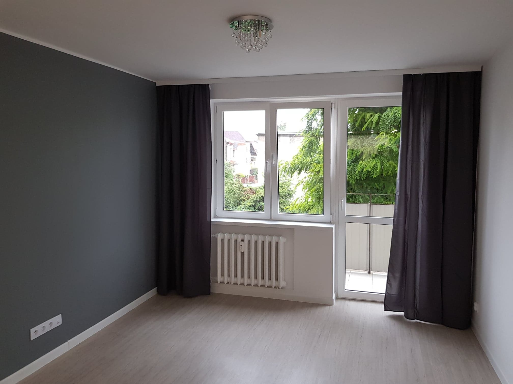 Pusty pokój po remoncie z szarą ścianą, jasnymi panelami podłogowymi, białym grzejnikiem pod oknem balkonowym i ciemnymi zasłonami.