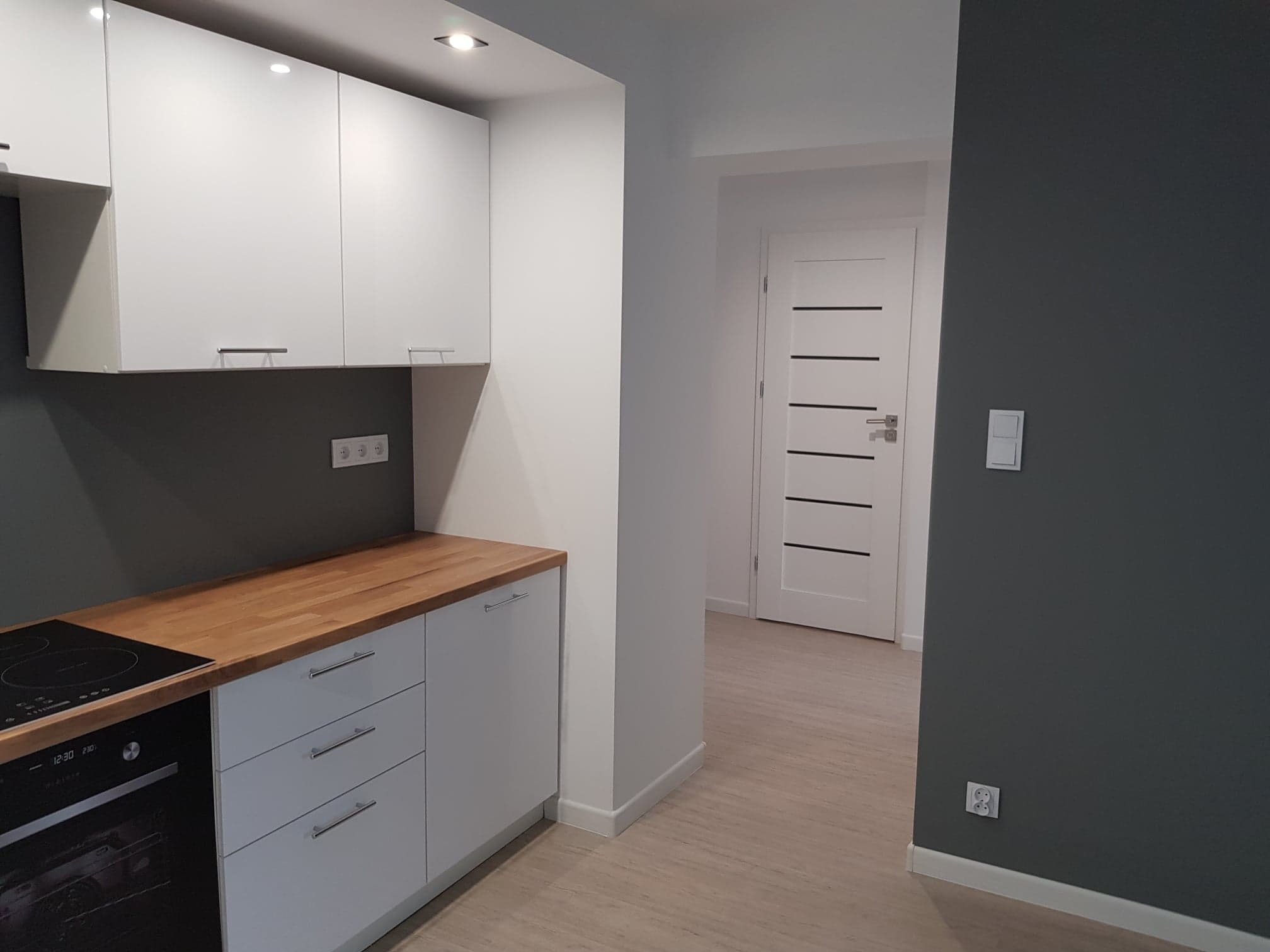 Nowoczesna kuchnia z białymi szafkami, drewnianym blatem i ciemnoszarą ścianą, widok na białe drzwi z poziomymi czarnymi paskami.