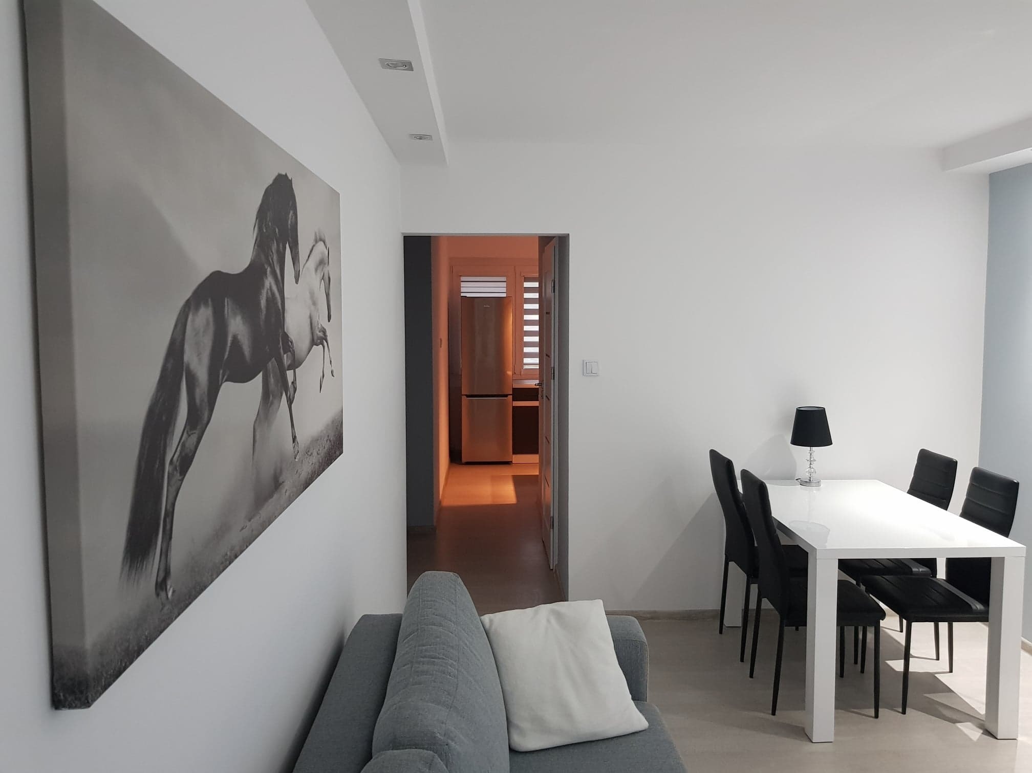 Jasne wnętrze mieszkania z szarą sofą, białym stołem z czarnymi krzesłami i obrazem z motywem koni na ścianie.