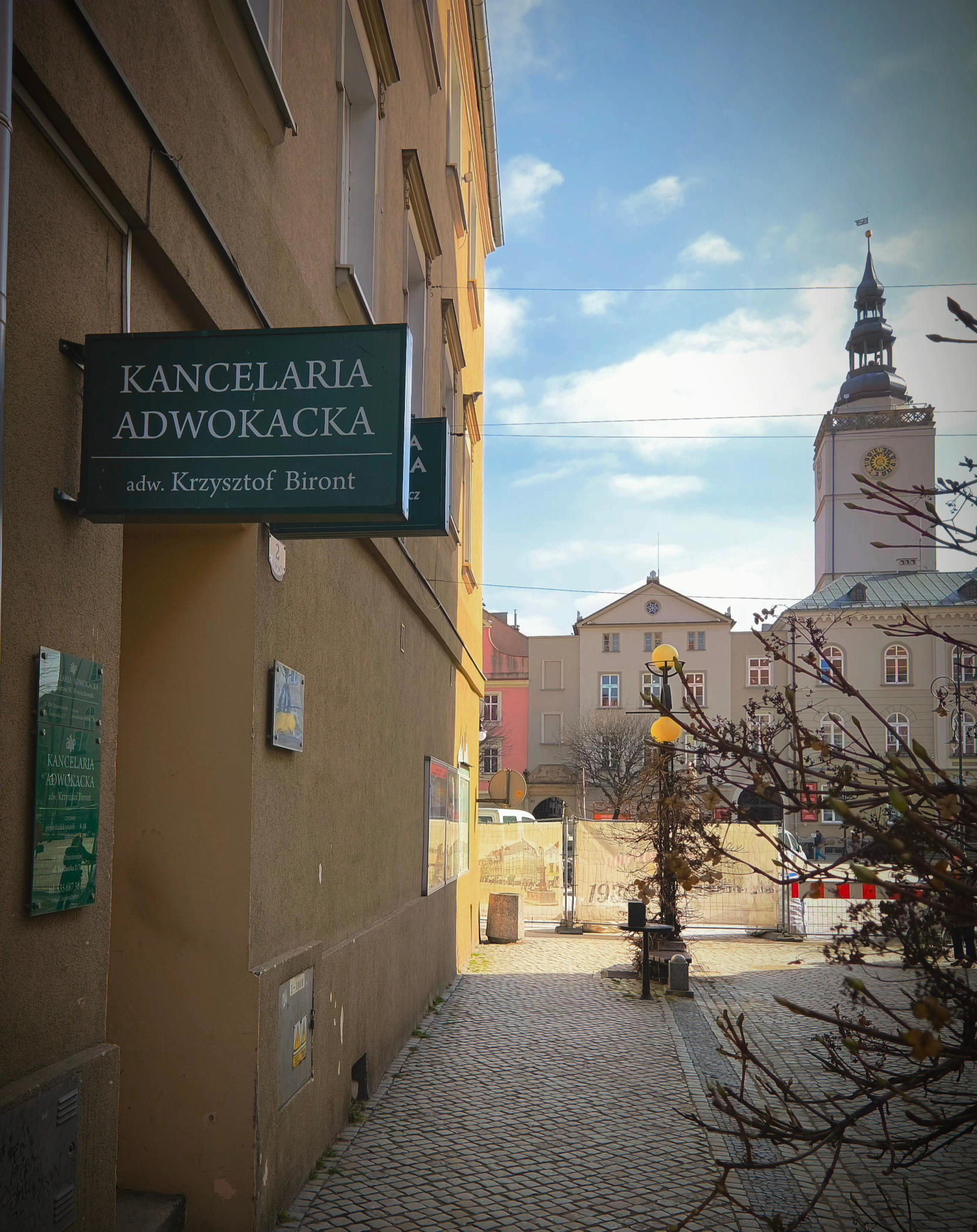 Zewnętrzny widok na budynek Kancelarii Adwokackiej adw. Krzysztofa Bironta z szyldem i tabliczką informacyjną, na tle budynku ratusza miejskiego i brukowanej uliczki.
