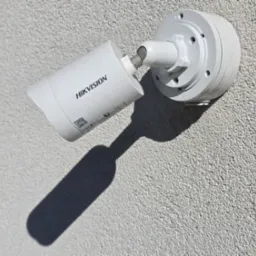 Kamera monitoringu Hikvision zamontowana na szarej, chropowatej ścianie budynku, rzucająca wyraźny cień. Widoczne śruby montażowe i etykieta z kodem QR.