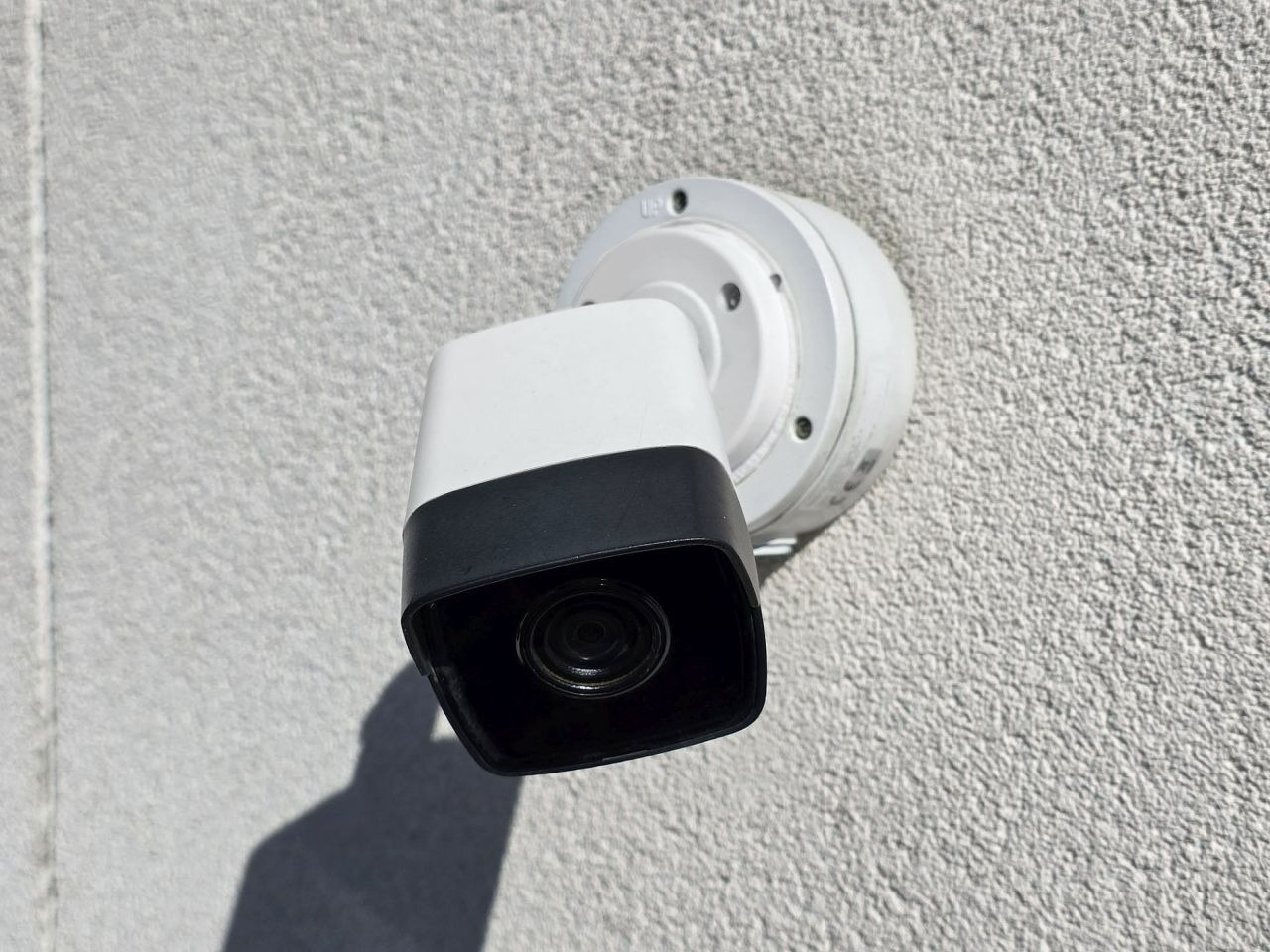 Kamera monitoringu (biało-czarna) zamontowana na szarej, chropowatej ścianie budynku. Widoczny cień kamery. Kąt ujęcia z dołu, światło dzienne.
