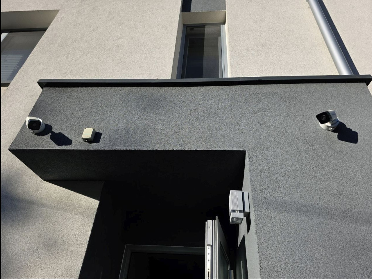 Dwie kamery monitoringu zamontowane na elewacji budynku w kolorze szarości. Dodatkowo widoczny czujnik alarmowy i oświetlenie zewnętrzne nad drzwiami.