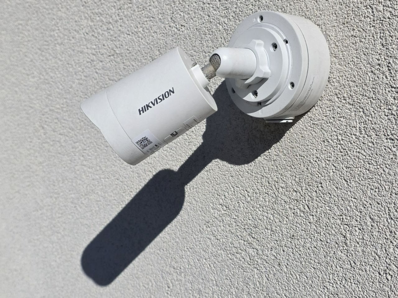 Kamera monitoringu Hikvision zamontowana na szarej, chropowatej ścianie budynku, rzucająca wyraźny cień. Widoczne śruby montażowe i etykieta z kodem QR.