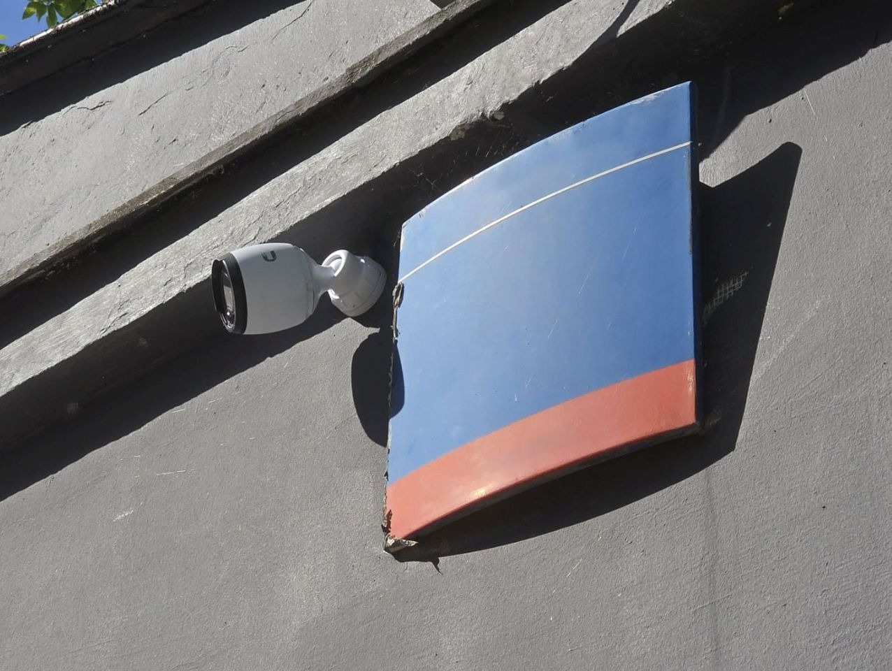 Kamera monitoringu IP zamontowana na szarej ścianie obok niebiesko-czerwonego szyldu, Warszawa. Widoczne okablowanie. Instalacja systemów zabezpieczeń.