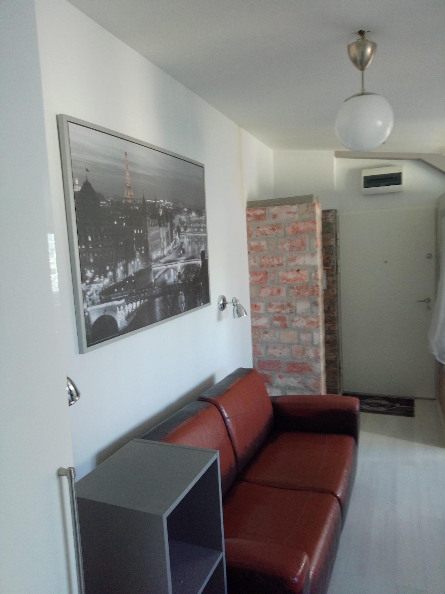 Wnętrze pokoju z czerwoną skórzaną sofą, szarą szafką, obrazem z wieżą Eiffla na ścianie, lampą sufitową w kształcie kuli i fragmentem ściany z czerwonej cegły.