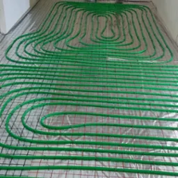 Rozłożona instalacja ogrzewania podłogowego z zielonych rurek na folii z siatką, widoczne okno i ściany w trakcie wykańczania.