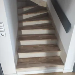 Schody wewnętrzne z drewnianymi stopniami, szare malowane ściany i czarna poręcz, widok z dołu, gniazdko elektryczne na ścianie po lewej stronie.