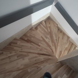 USŁUGI BUDOWLANE ROMAN BORUKH - Spiralne schody drewniane z jasnego drewna, widok z góry, fragment stopy w klapku na dole kadru, ciemne listwy przypodłogowe.