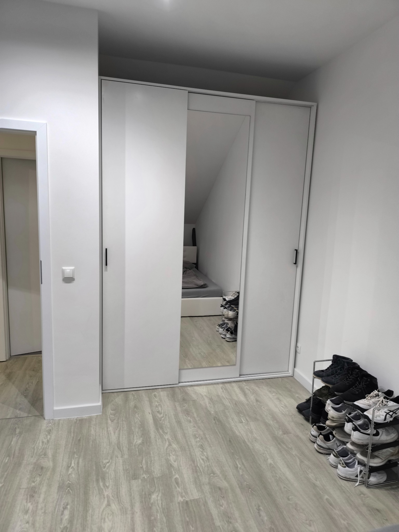 Wnętrze pokoju z białą szafą przesuwną z lustrem, odbijającym skośny sufit i łóżko, oraz półką na buty w rogu.