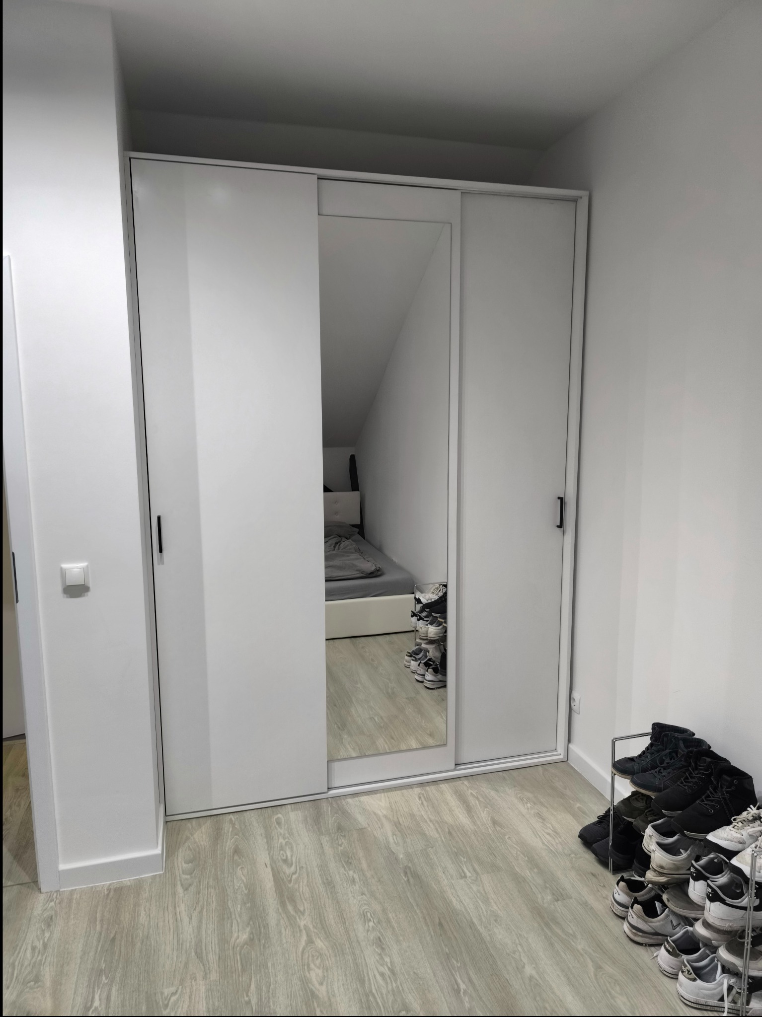 Biała szafa przesuwna z lustrem w pokoju z poddaszem; widoczne półki na buty i fragment łóżka odbijający się w lustrze.