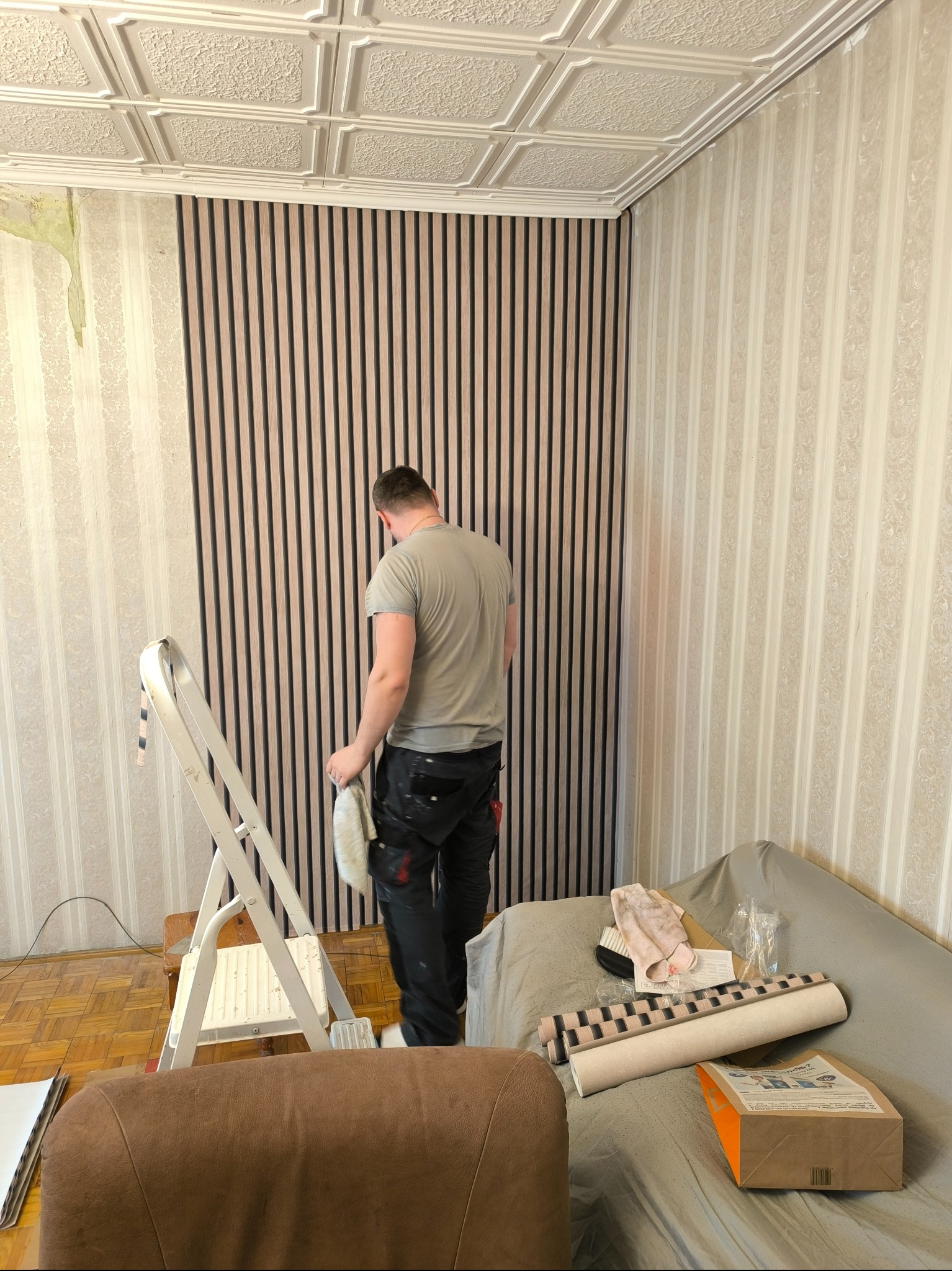 Mężczyzna w trakcie montażu paneli ściennych z pionowymi listwami na tle tapety w paski, widoczna drabina i materiały budowlane na sofie.