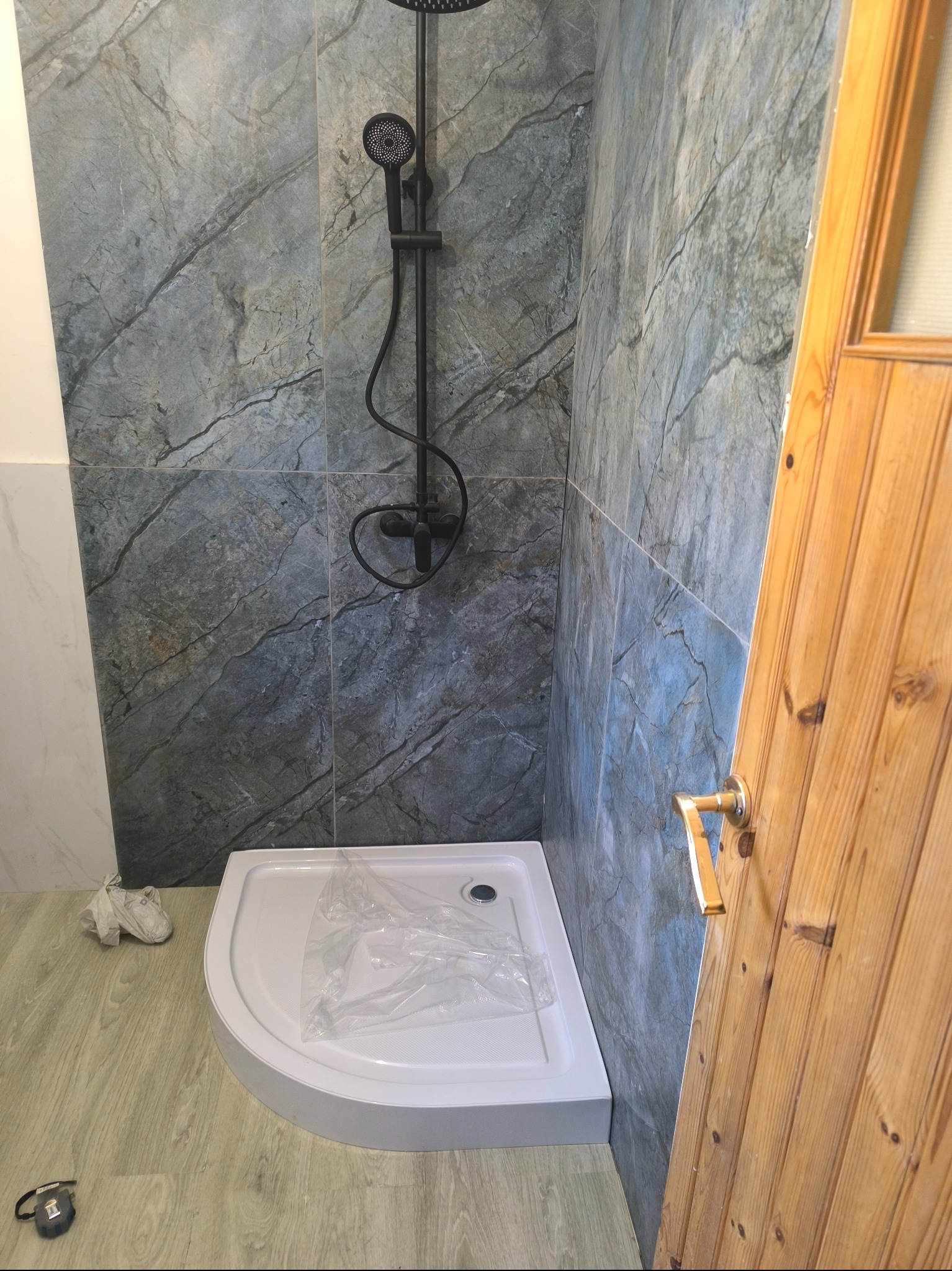 Narożny brodzik z czarną armaturą prysznicową na tle ściany wyłożonej szarymi płytkami imitującymi marmur, widoczna drewniana ościeżnica drzwi i podłoga z panelami.