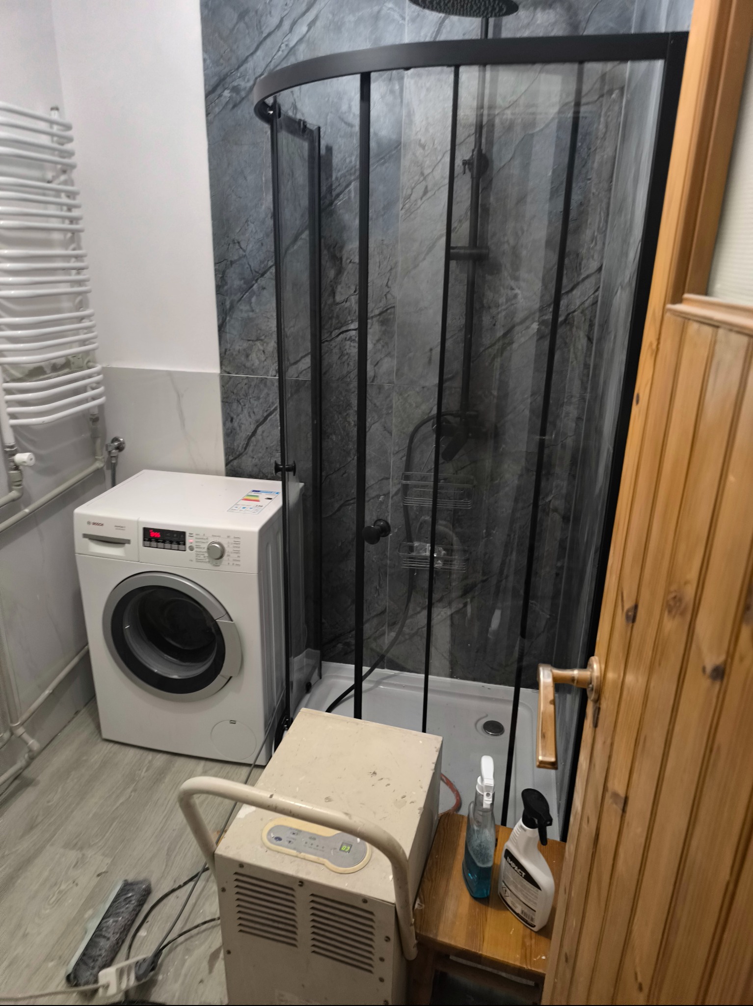 Łazienka po remoncie: kabina prysznicowa z czarnymi ramami, pralka, osuszacz powietrza i drewniane drzwi. Widoczne butelki ze środkami czystości.