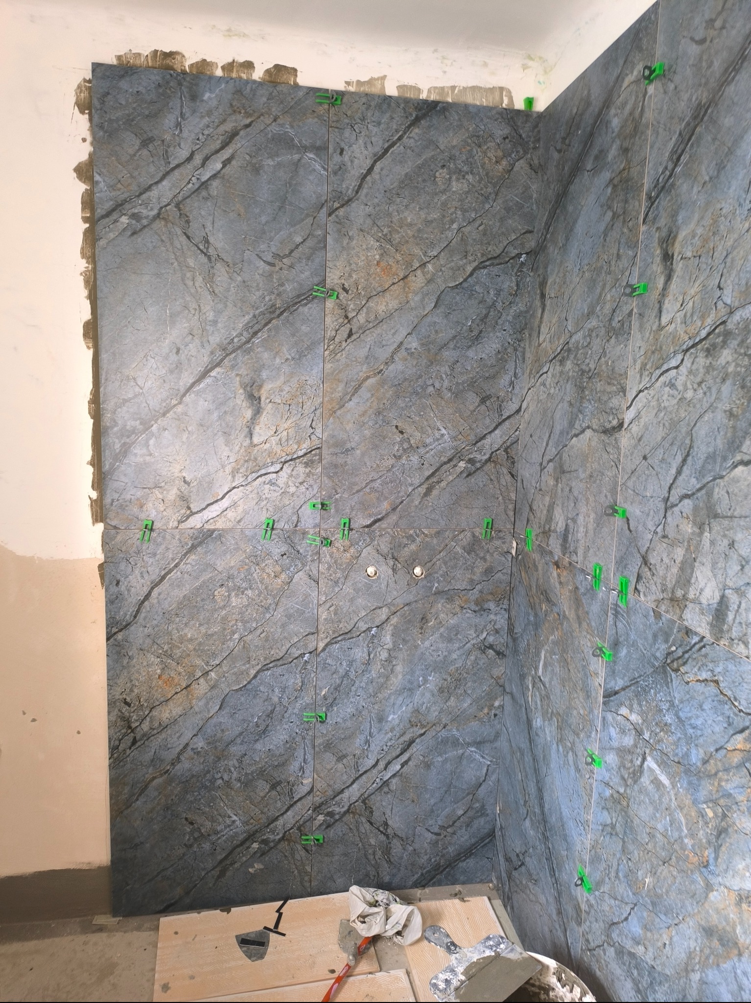 Płytki imitujące marmur na ścianie łazienki w trakcie montażu, z użyciem zielonych klipsów poziomujących, widoczne narzędzia i resztki kleju na podłodze.